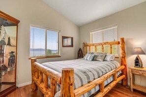 Un dormitorio con estructura de cama de madera, un ventanal con persianas, una mesita de noche con lámpara, y un cuadro enmarcado en la pared.