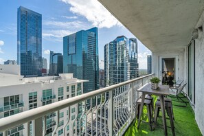 A balcony with a view of tall buildings, a table with chairs, and a small indoor space with a door.