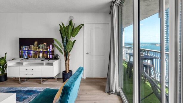 Un salon moderne avec vue sur la ville, un meuble de rangement blanc pour les appareils électroniques, une grande plante en pot et un balcon doté d’une balustrade.