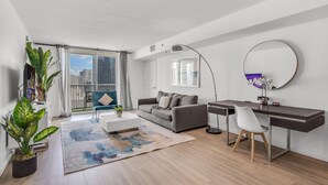 Un salon moderne avec un canapé gris, une table basse blanche, un bureau avec une chaise, une grande fenêtre avec des rideaux et un balcon offrant une vue sur les immebles.