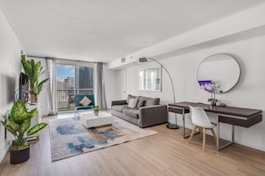 A modern living room with a grey sofa, a white coffee table, a desk with a chair, a large window with curtains, and a balcony with a view of buildings.