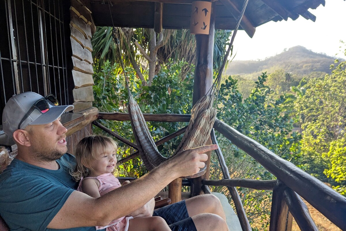 A man and a child sitting on a wooden bench under a wooden structure, with a view of greenery and hills in the background.