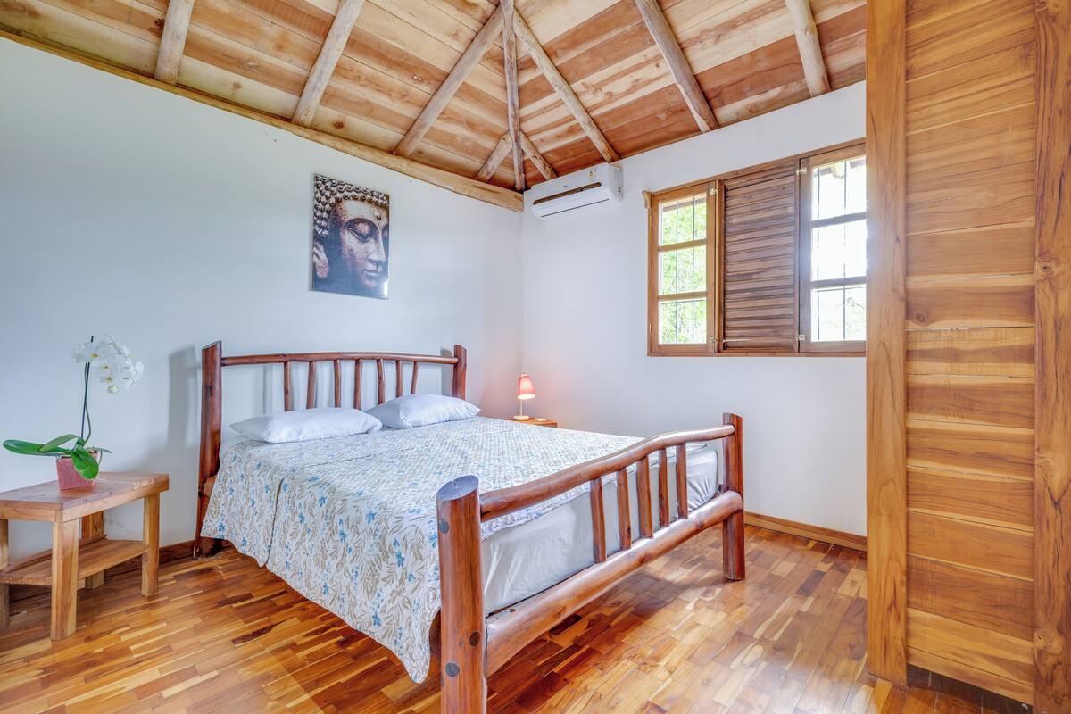 A wooden-framed bed with a patterned bedspread, a nightstand with a lamp, a wall-mounted air conditioner, and a window with wooden shutters.