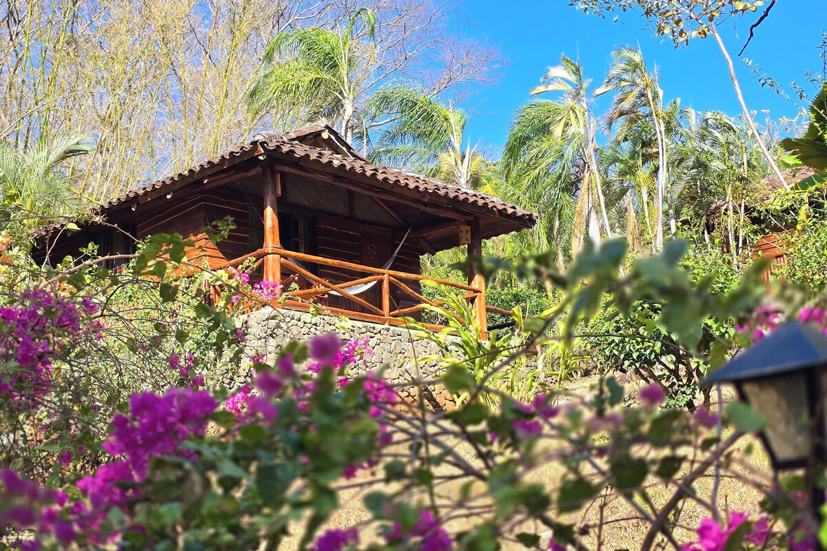 A rustic wooden cabin with a stone foundation, surrounded by lush greenery and flowering plants.