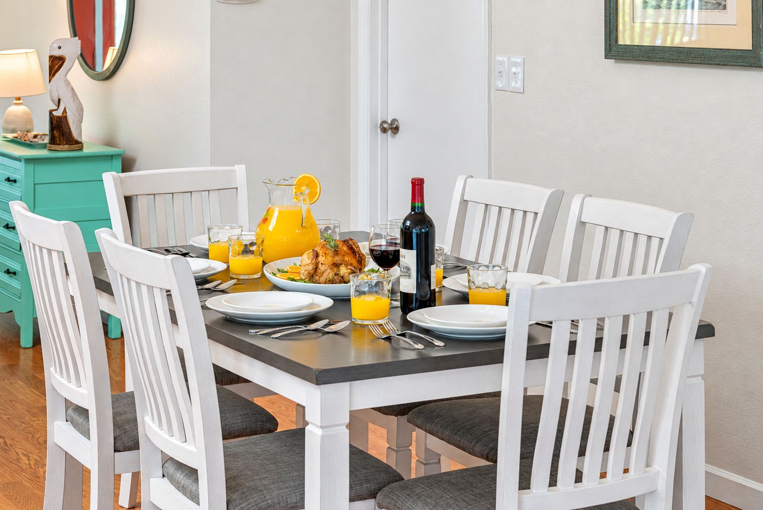 Mesa de comedor lista para seis personas, con sillas blancas, una mesita auxiliar color turquesa y un frutero.