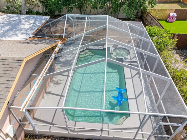 A swimming pool with a glass enclosure, surrounded by a wooden deck and a fenced area with a small pink structure.