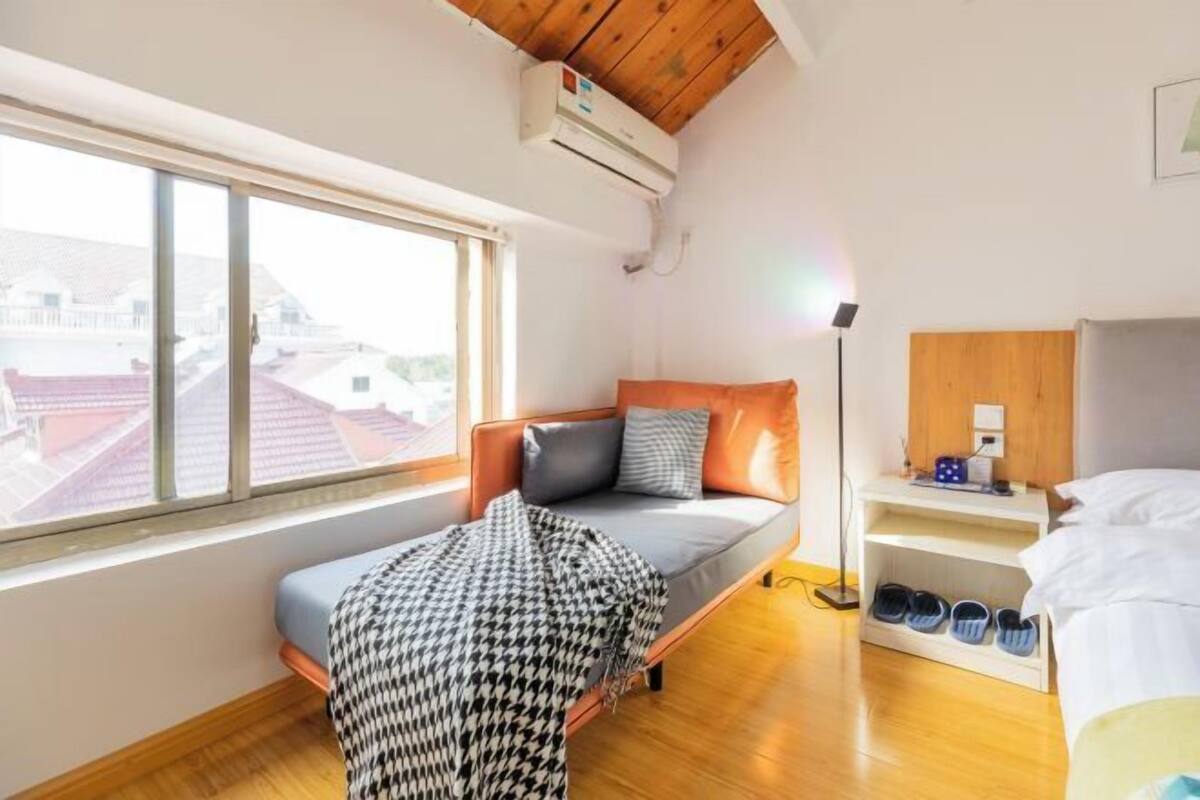 A bedroom with a bed, a bedside table, a shelf with shoes, a window, and an air conditioning unit.