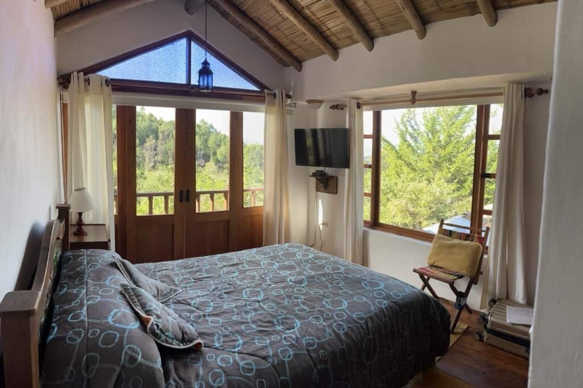 Un dormitorio con una cama grande, un televisor montado en la pared y vistas al exterior a través de amplios ventanales.