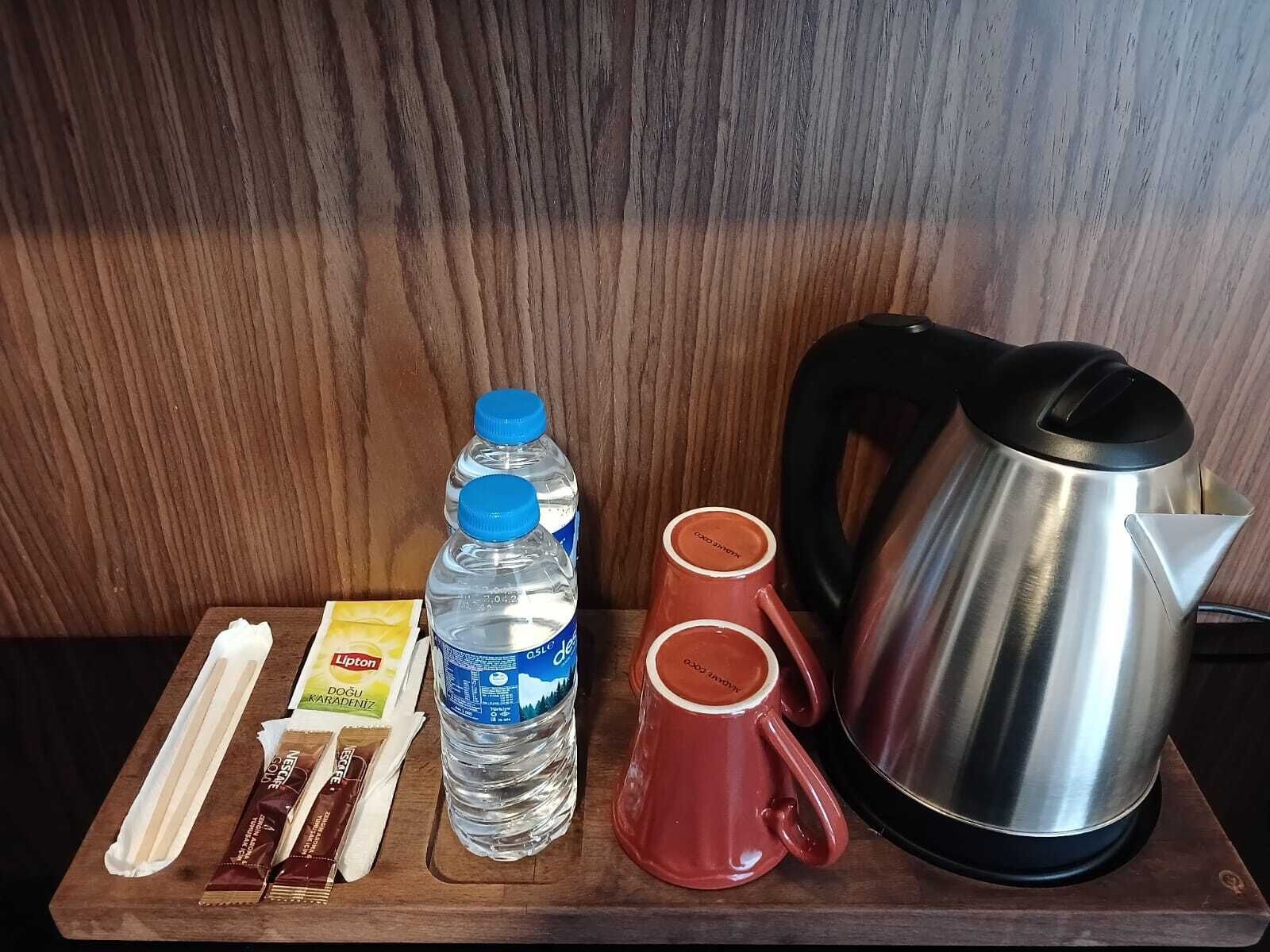 Una bandeja de madera con un hervidor de acero inoxidable, dos tazas rojas, una botella de agua, un sobre de té Lipton y dos barras de chocolate.
