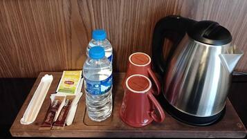 Una bandeja de madera con un hervidor de acero inoxidable, dos tazas rojas, una botella de agua, un sobre de té Lipton y dos barras de chocolate.