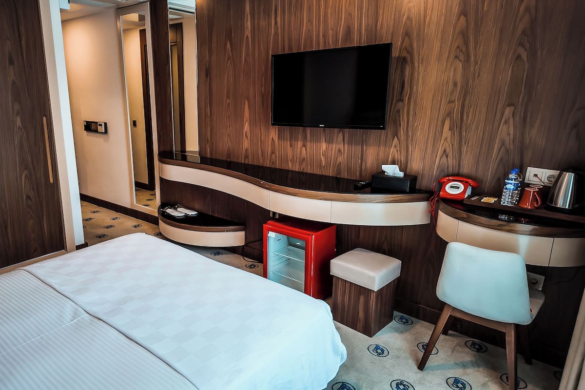 Una habitación de hotel con una cama, un escritorio con espejo, un televisor, un teléfono rojo y un refrigerador pequeño.