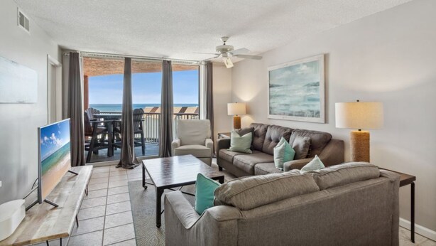 A living room with a sofa, armchairs, a coffee table, and a TV. There is a balcony with a view of the ocean.