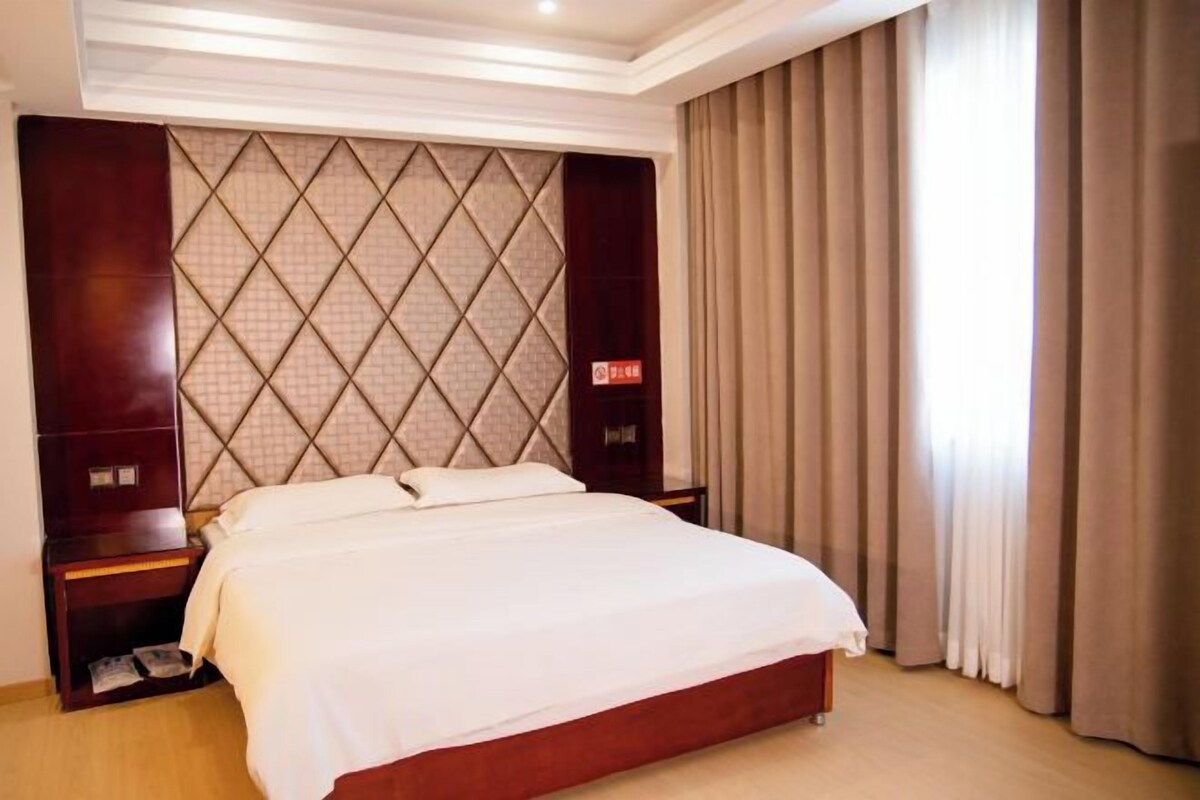 A hotel room with a large bed, a headboard with a diamond pattern, a nightstand, and a window with curtains.