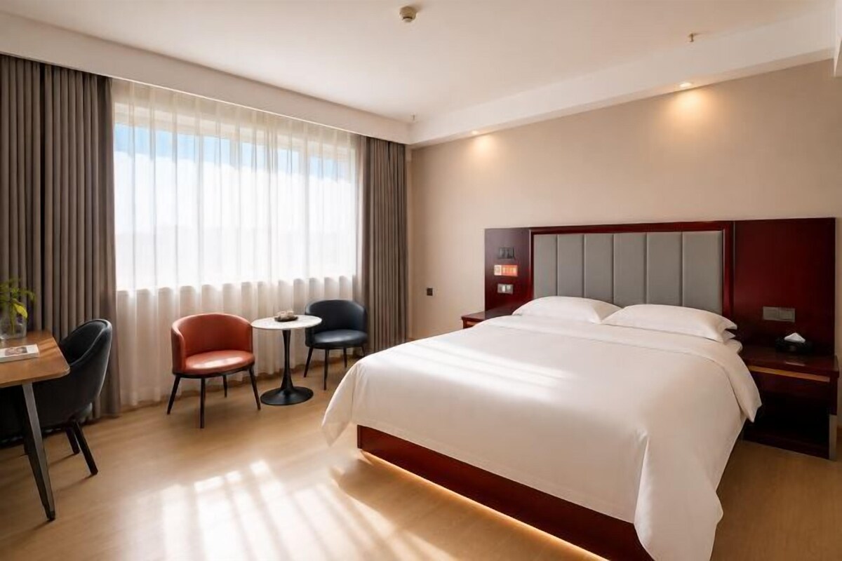 A hotel room with a large bed, a desk with two chairs, a small round table, and a nightstand with a lamp.