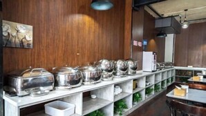 Un espace buffet avec des plats de service en acier inoxydable disposés sur des étagères, un mur en bois orné d’un tableau représentant des fleurs et une table avec des chaises.