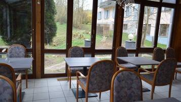 Ein Essbereich mit Tischen und Stühlen, große Fenster mit Blick auf einen Garten und Gebäude.