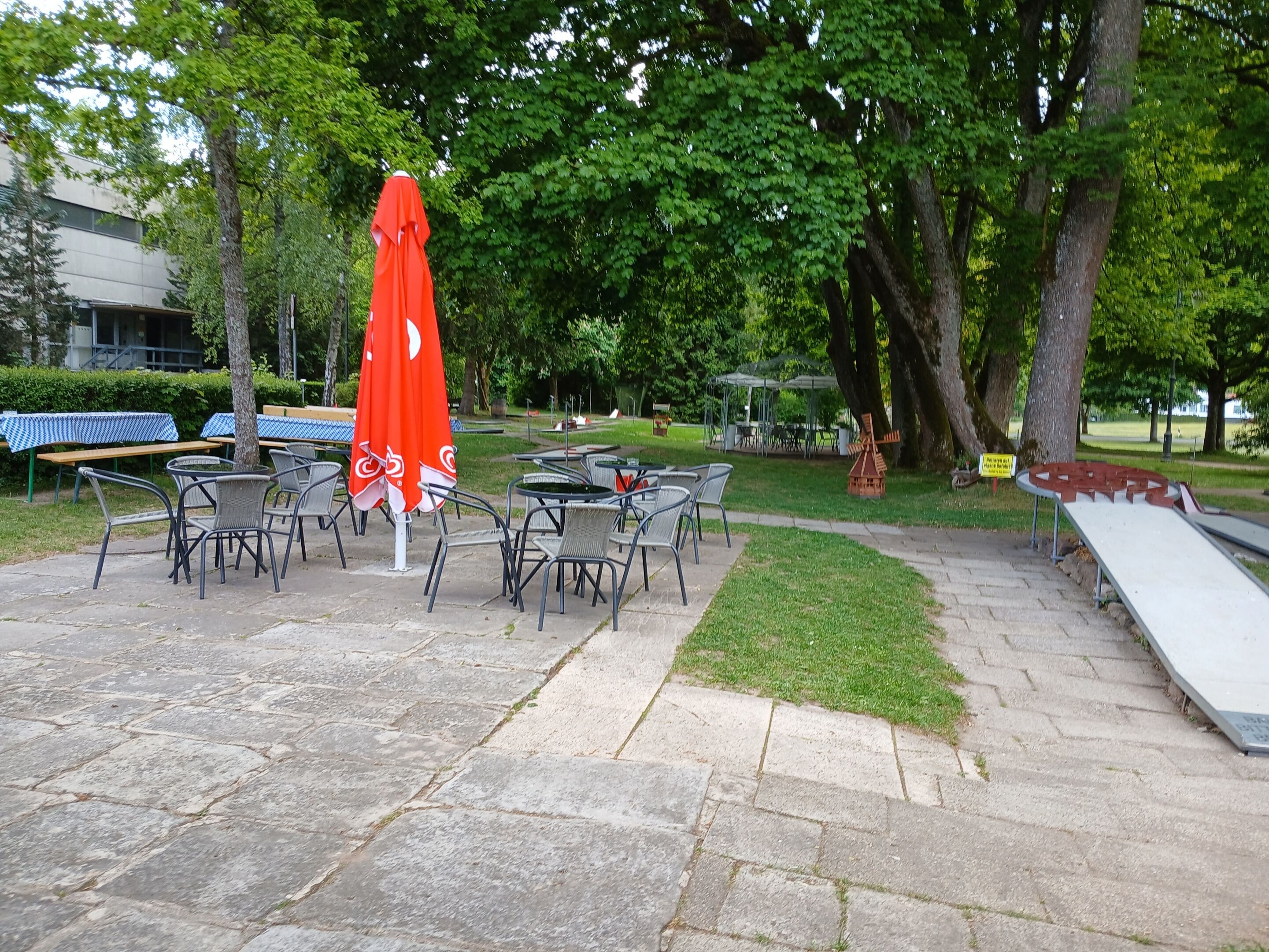 An outdoor seating area with tables and chairs, a red umbrella, and a playground.