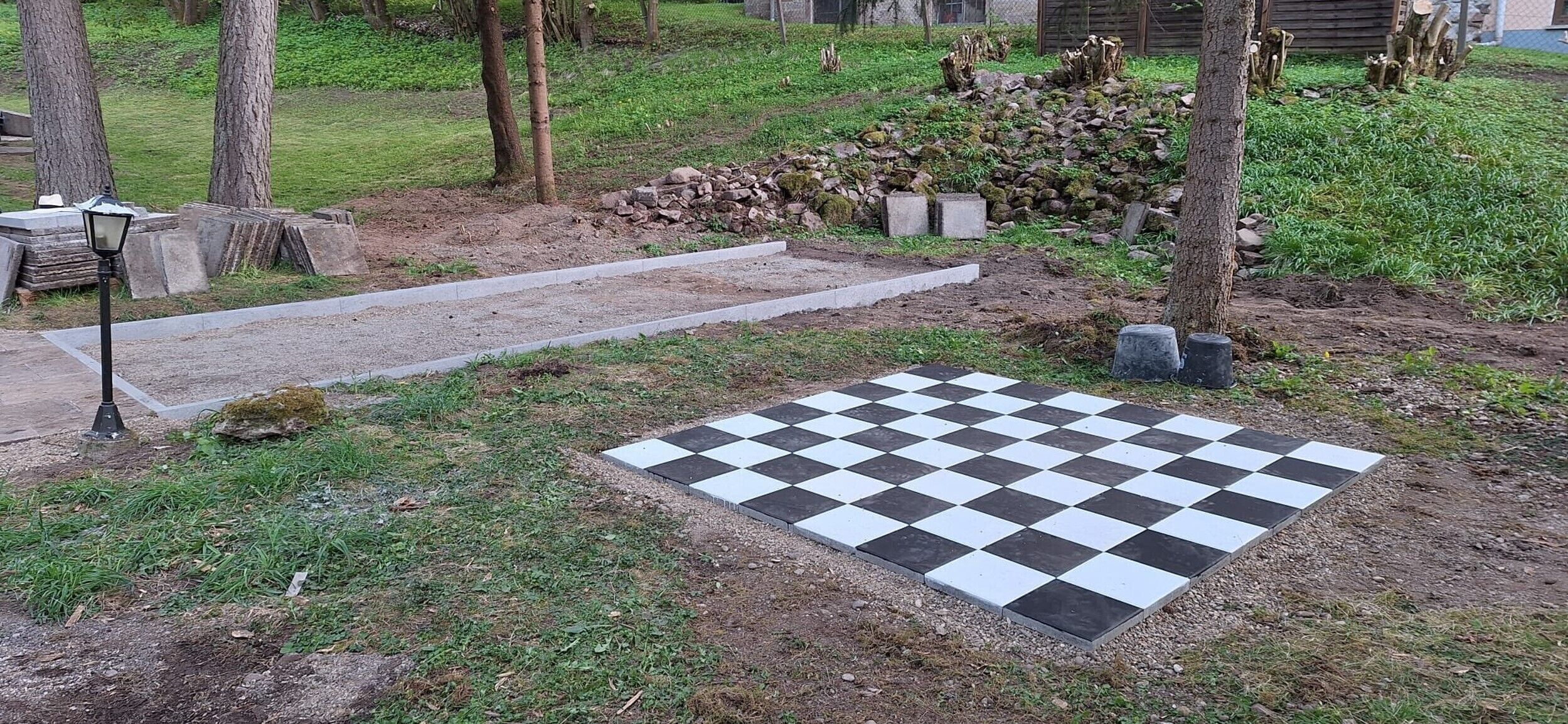 Un espace extérieur à motifs géométriques, avec un chemin pavé et des marches en pierre, entouré d’arbres et d’une pile de bûches.