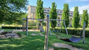 A playground with swings and a climbing structure, surrounded by grass and trees, with a building in the background. - Hotel am Park (Bad Brueckenau)