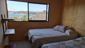 A small, cozy room with two beds, a wooden shelf, and a large window offering a view of a cityscape. - Ocean view in Rukalafquen (Dichato)