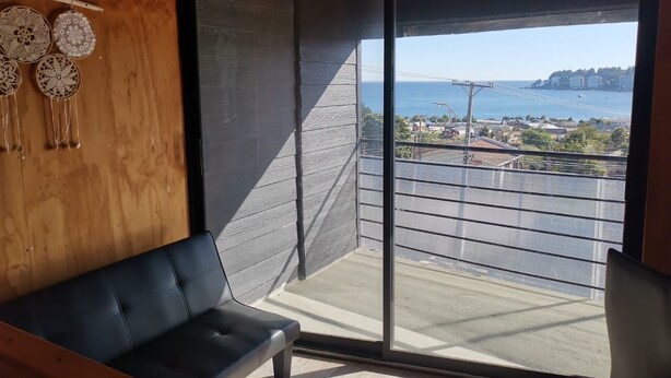 A modern room with a large sliding glass door leading to a balcony with a view of the sea and a bridge. - Ocean view in Rukalafquen (Dichato)