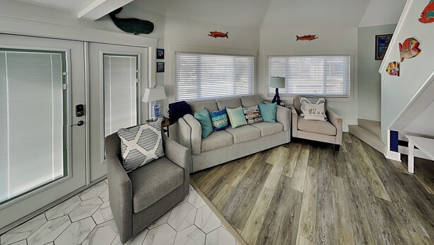 A modern living room with a sectional sofa, a small armchair, and a coffee table. There are two windows with blinds, a lamp, and decorative fish wall art. - Sunday Villas Sunday Funday by Silver Sands Vacation Rentals (Port Aransas)