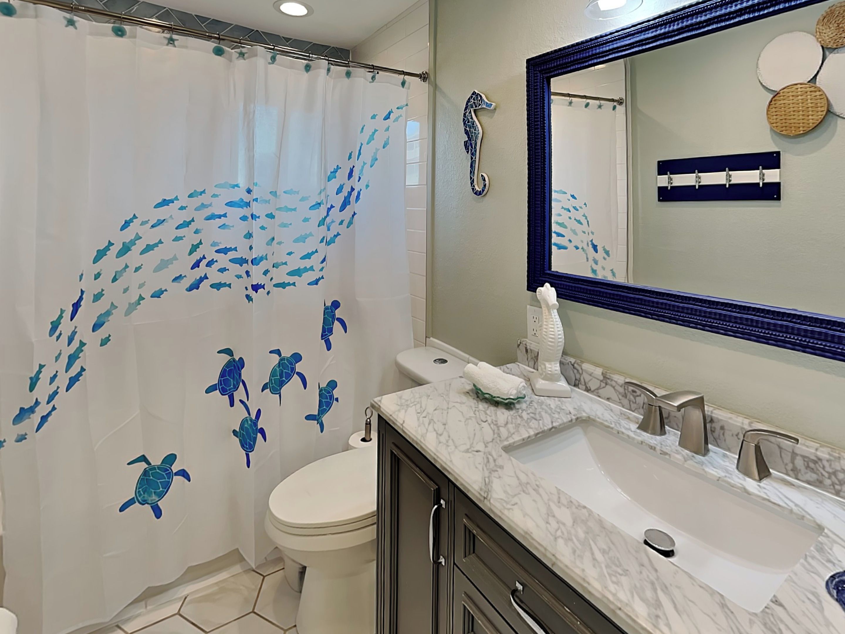 Une salle de bain avec un rideau de douche orné de motifs d’animaux marins, des toilettes blanches, un lavabo en marbre et un grand miroir.