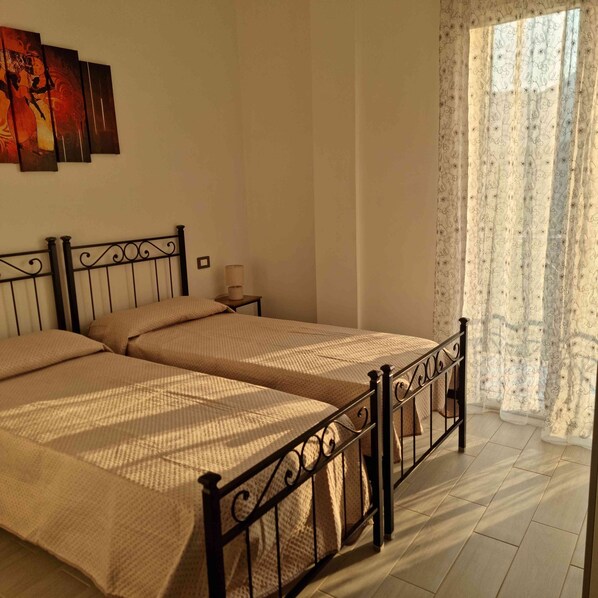 A double bed with a metal frame, a bedside table with a lamp, and a window with patterned curtains.