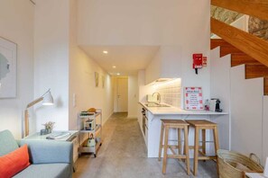 Ein kompakter Wohnraum mit Küchenzeile, einer Couch, einem Schreibtisch mit Lampe und einer Treppe.