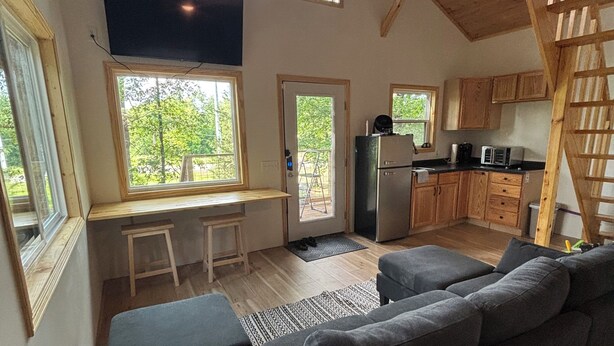 A cozy cabin interior with a large gray sofa, wooden floors, a kitchen area, and a ceiling fan.