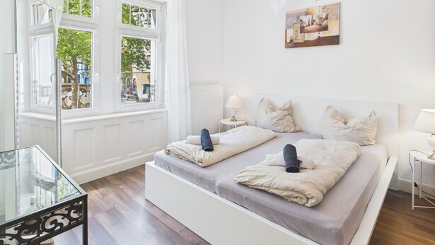 Un dormitorio moderno con una cama grande, mesitas de noche, una mesa de vidrio y vistas a la calle a través de un ventanal.