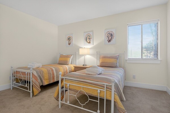 A bedroom with two beds, each with striped bedding and pillows, a bedside table with a lamp, and a window with blinds.