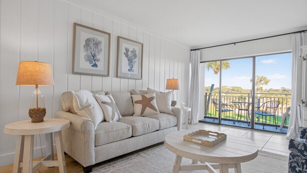 A modern living room with a sofa, two side tables, and a coffee table. There are framed botanical prints on the wall, and a balcony with outdoor furniture is visible. - 2BR Golf View Villa Near Beach in Sea Pines Resort (Hilton Head Island)