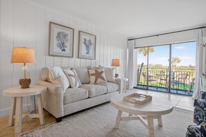A modern living room with a sofa, two side tables, and a coffee table. There are framed botanical prints on the wall, and a balcony with outdoor furniture is visible.