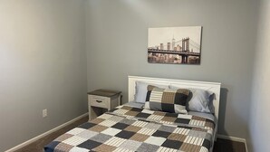 Quarto com colcha xadrez, cabeceira branca, mesa de cabeceira e quadro com vista para a cidade.