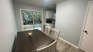 Uma sala de jantar com mesa e cadeiras de madeira, uma vela acesa e vista para a cozinha e áreas verdes ao ar livre.