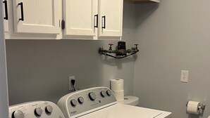 Uma área de lavanderia com máquina de lavar e secadora frontais, armários brancos acima e um vaso sanitário com suporte para toalhas de papel.