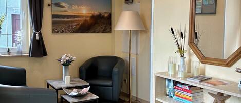 A living room with a sofa, a chair, a table with a vase, a lamp, a bookshelf, and a painting on the wall.