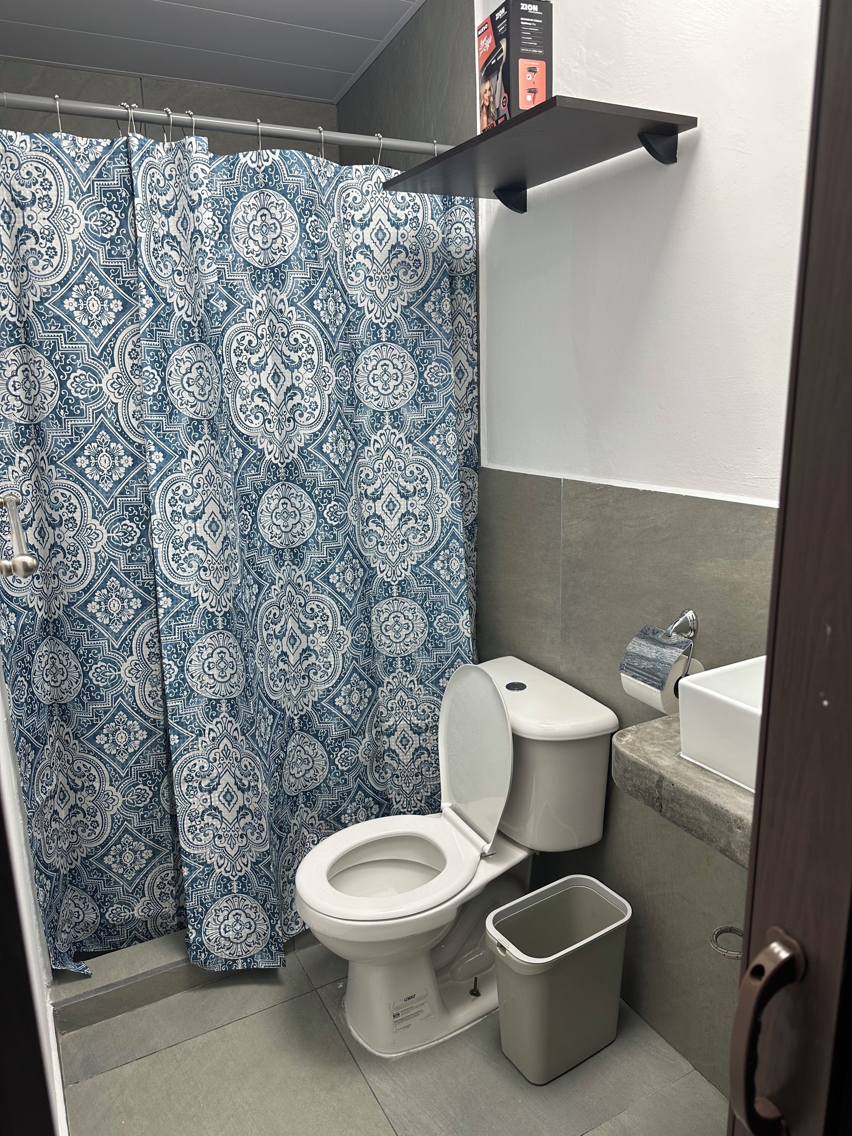 Ein Badezimmer mit gemustertem Duschvorhang, Toilette, Mülleimer und Ablage mit einer Schachtel.