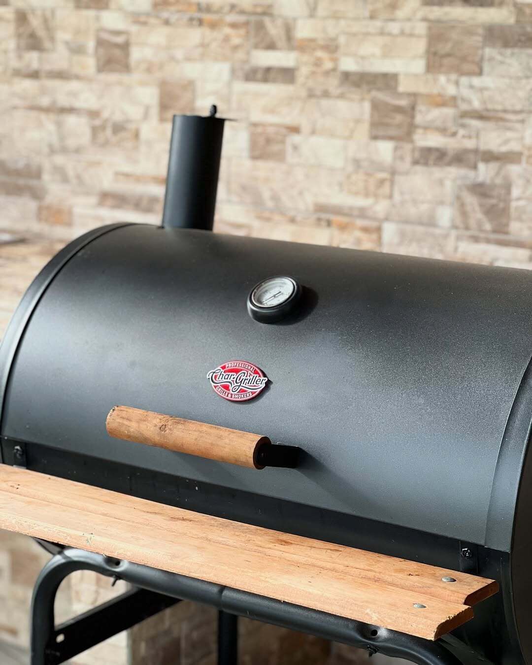 A charcoal grill with a wooden handle and a pressure gauge, set against a stone wall background.