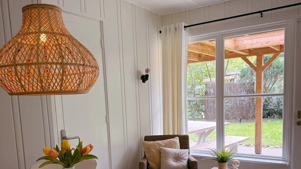 Un coin repas avec une table ronde, des chaises, un luminaire suspendu en osier, un vase avec des fleurs et une vue sur l’extérieur par le biais d’une grande fenêtre.