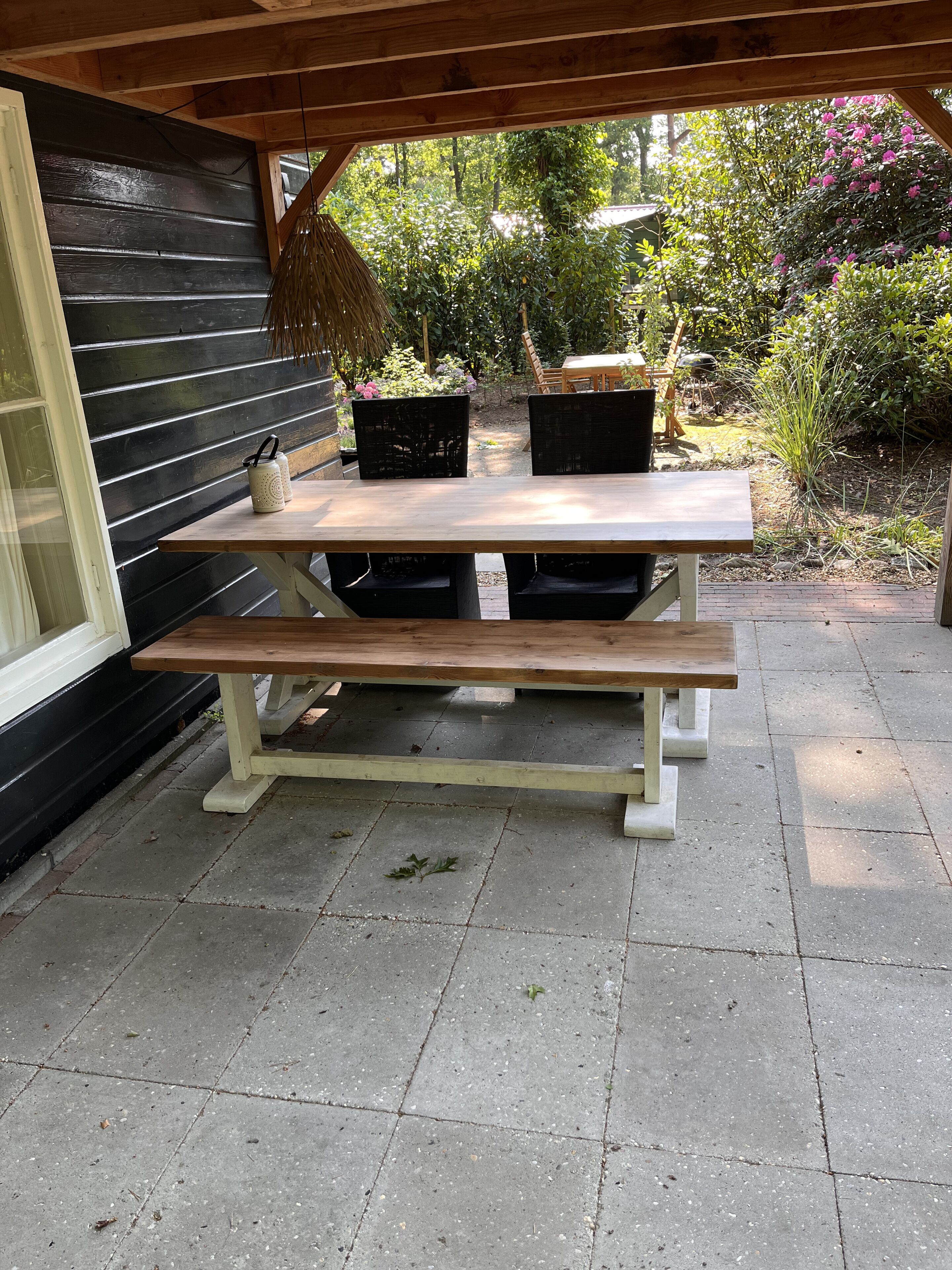 A wooden picnic table with benches, set on a paved patio with a garden in the background.
