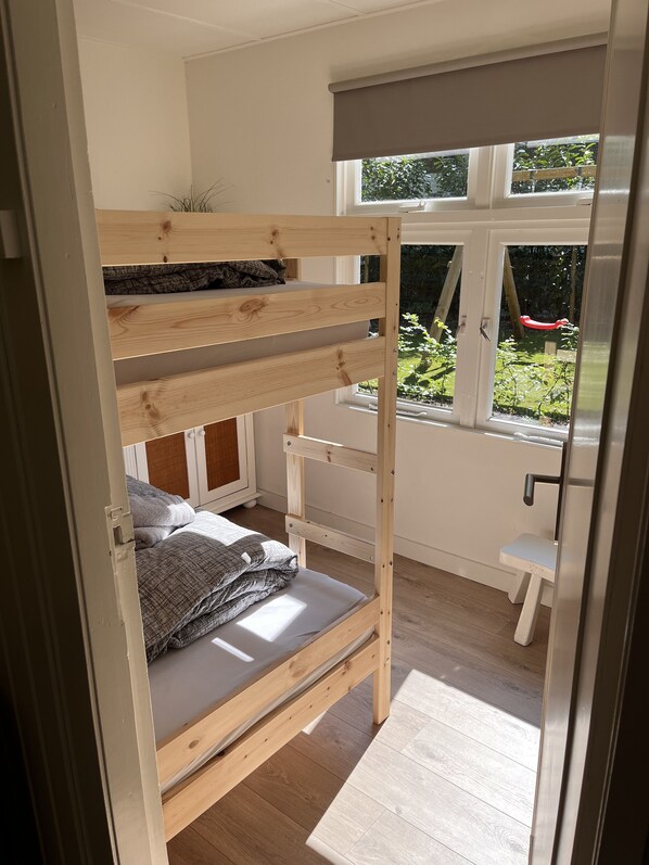 A bunk bed with a lower bunk and a small desk, a window with a view of greenery, and a wooden floor.