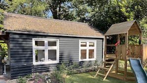 Une maison avec deux fenêtres, une cabane en bois avec un toboggan, et un jardin avec des plantes et une pelouse.