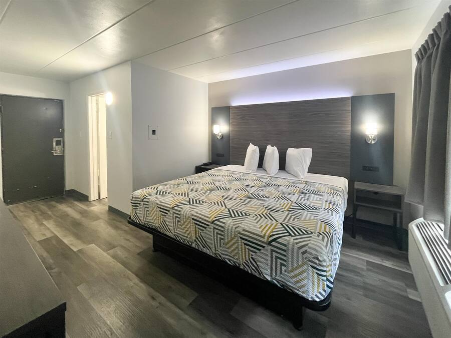 A modern hotel room with a large bed, a nightstand, a wall-mounted lamp, and a door.