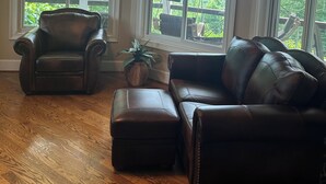 A spacious living room with a leather sofa, a leather armchair, and a wooden floor. Large windows offer a view of the outdoors.