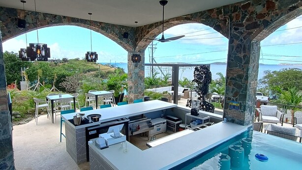 An outdoor pool area with a bar and seating, surrounded by stone arches and a view of the sea. - The Saint Resort (St. John)