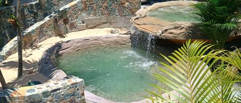 Un área de aguas termales con paredes de piedra y una pequeña piscina, rodeada de abundante vegetación.