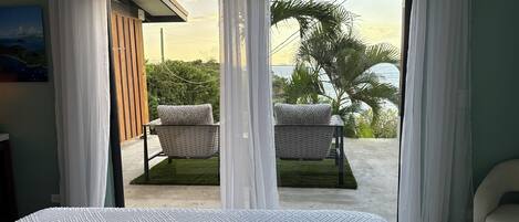 A bedroom with a bed, two wicker chairs with cushions, a view of a balcony with greenery and a body of water.