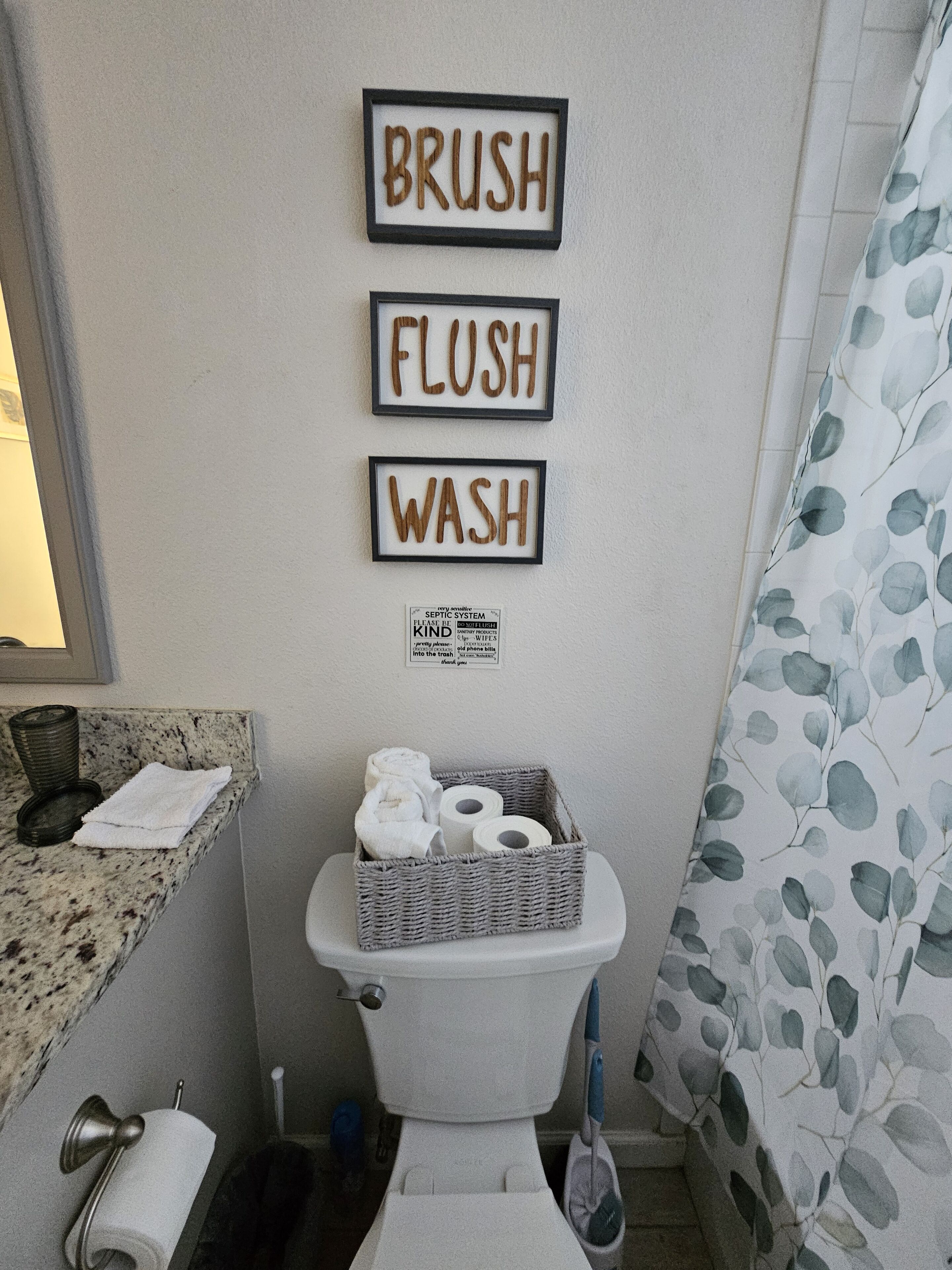 Une salle de bain avec des toilettes, un panier contenant des articles de toilette et trois mots encadrés accrochés au mur : « BRUSH », « FLUSH », « WASH ».